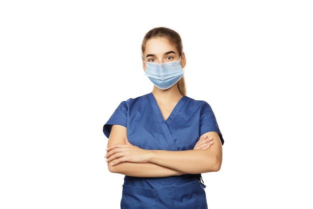 Young beautiful female doctor in blue surgical gown and medical disposable mask on face with folded hands on chest isolated on white