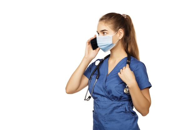 Young beautiful female doctor in blue surgical gown with stethoscope speaks on the phone with a patient isolated on white background Copspace