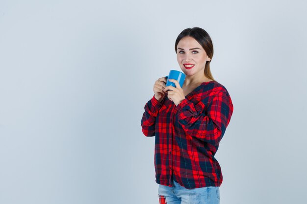 Young beautiful female holding cup near mouth in casual shirt, jeans and looking joyful , front view.