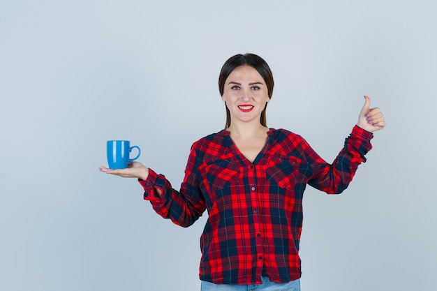 Giovane bella tazza di contenimento femminile mentre mostra pollice in su in camicia casual, jeans e sembra gioiosa. vista frontale.