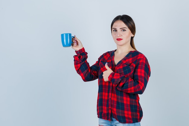 Молодая красивая женщина держит чашку, показывая большой палец вверх в повседневной рубашке, джинсах и выглядит оптимистично, вид спереди.