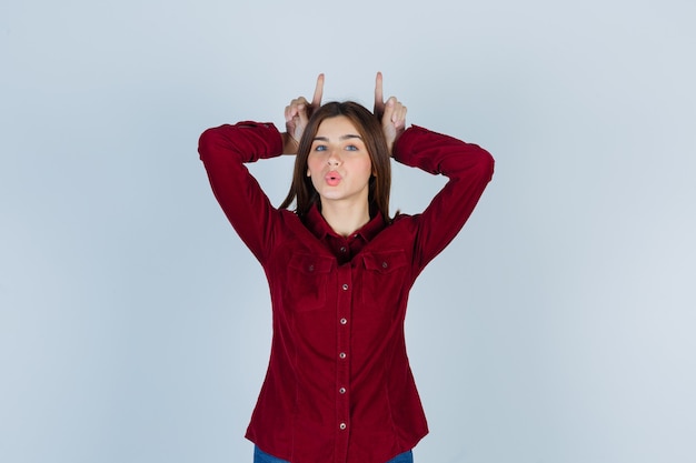 Young beautiful female keeping fingers over head as horns in shirt and looking playful.