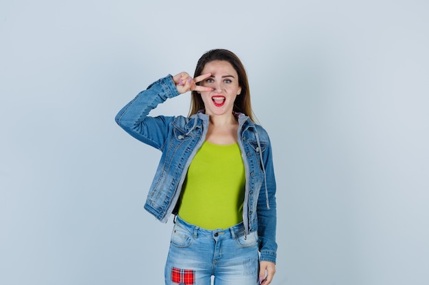 Young beautiful female looking through fingers in denim outfit and looking merry , front view.