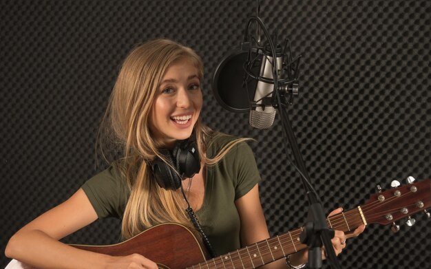 Young beautiful female musician with guitar is testing the\
sound system in the recording studio