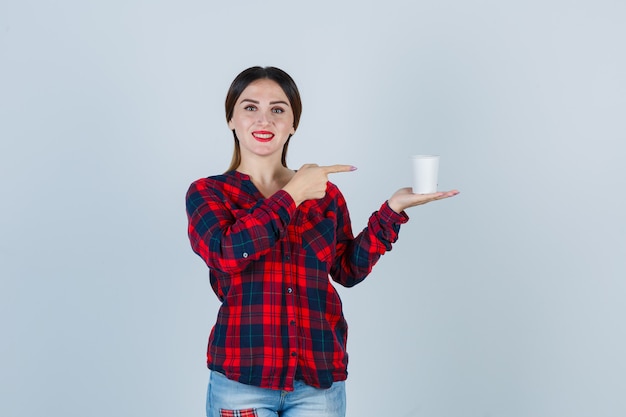 Giovane bella donna che punta al bicchiere di plastica in camicia casual, jeans e sembra allegra. vista frontale.