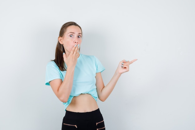 Young beautiful female pointing right, covering mouth with hands in t-shirt, pants and looking shocked. front view.