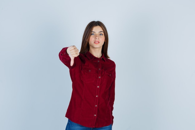 Young beautiful female showing thumb down in shirt and looking cheerless