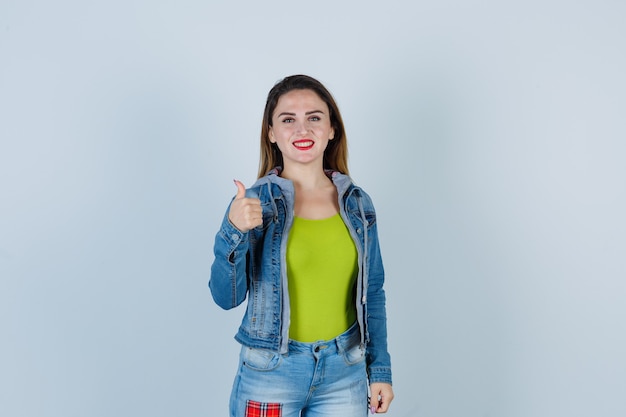 Young beautiful female showing thumb up in denim outfit and looking merry. front view.