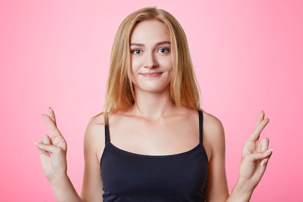 Young beautiful female student keeps fingers crossed, makes desired wish, believes in success or good luck