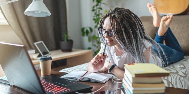 A young beautiful female student with dreadlocks is studying in an online lesson at home in a room with a laptop