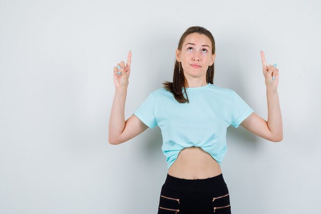 Young beautiful female in t-shirt, pants pointing up, looking upwards and looking blissful , front view.