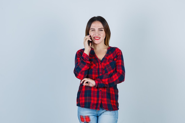 Young beautiful female talking on phone, winking eye in casual shirt and looking merry. front view.