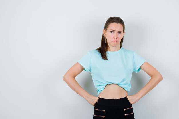 Young beautiful female with hands on waist in t-shirt, pants and looking grumpy. front view.