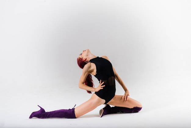 Young beautiful flexible female in a black jumpsuit and high heels is posing in a dance studio.