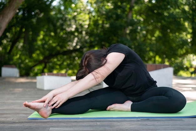 Young beautiful flexible girl makes a lean forward with her hands from a sitting position. Yoga class in the morning in the park. Side view