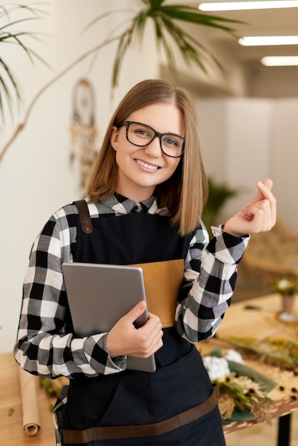 Young beautiful florist woman holding tablet and showing symbol of love with her fingers