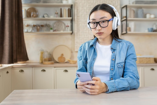 Young beautiful focused asian woman listening to audiobook podcast sitting at home in the kitchen