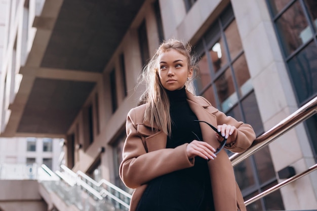 A young beautiful girl in a beige coat with sunglasses stands on the street. clothing advertisement