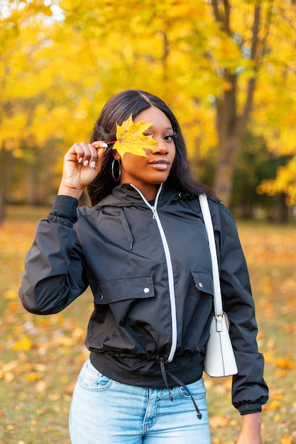 Young beautiful girl in a black casual jacket and blue jeans with a white handbag covering her face with a yellow autumn leaf on golden nature outdoors