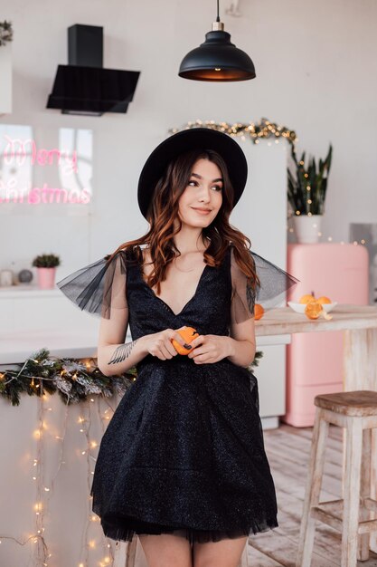 Young beautiful girl in a black dress and a hat cleans a tangerine