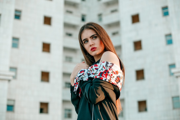 Young beautiful girl in a black jacket with a scarf poses near a high white house.