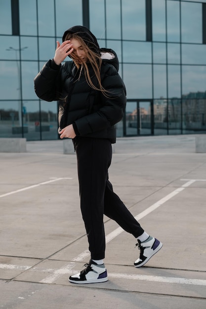 A young beautiful girl in a black short down jacket and sweatpants stands near a mirror wall Long haired woman stands near a building and looks down