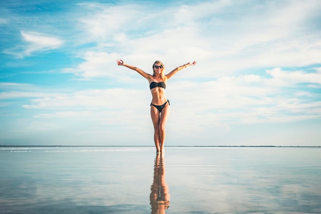 A young beautiful girl in a black swimsuit is rejoicing by raising her hands up against the background of a blue sky with clouds and clear water in salt lake The concept of vacation and travel