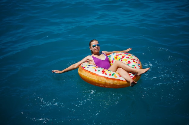 A young beautiful girl in a bright swimsuit lies on a large inflatable ring and floats on the blue sea on a bright sunny summer day