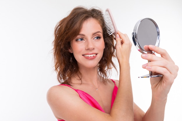 Young beautiful girl brushes her hair with little mirror isolated on white wall