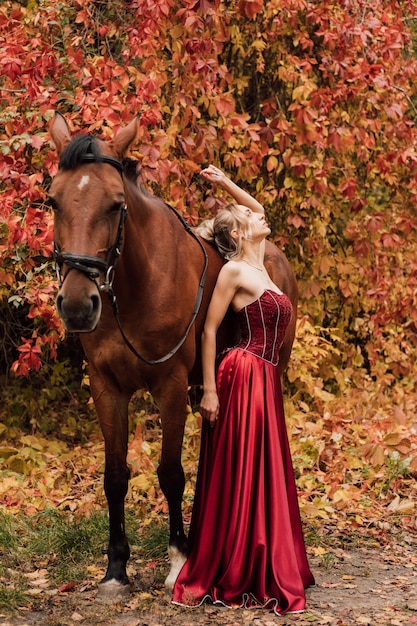 Young beautiful girl in a burgundy long dress walks with a\
horse in the autumn forest