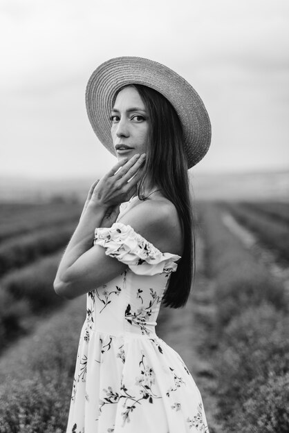 A young beautiful girl in a delicate dress and hat walks through a beautiful field of lavender and enjoys the aroma of flowers. Vacation and beautiful nature. Black and white.