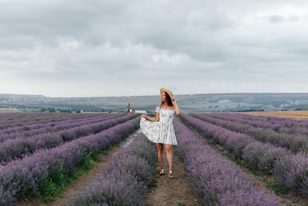 A young beautiful girl in a delicate dress and hat walks through a beautiful field of lavender and enjoys the fragrance of flowers Rest and beautiful nature Lavender blooming and flower picking