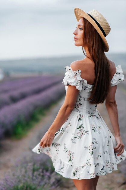 A young beautiful girl in a delicate dress and hat walks through a beautiful field of lavender and enjoys the fragrance of flowers Rest and beautiful nature Lavender blooming and flower picking