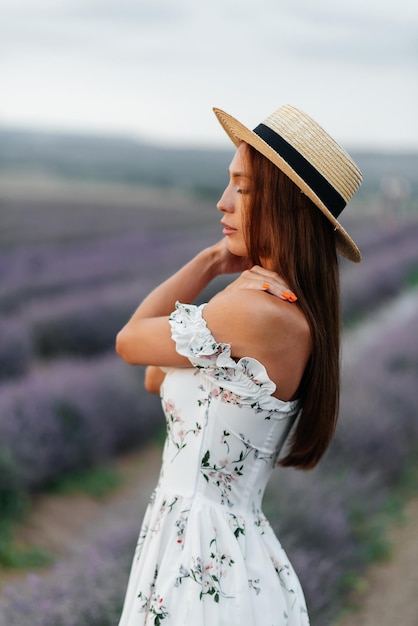 A young beautiful girl in a delicate dress and hat walks\
through a beautiful field of lavender and enjoys the fragrance of\
flowers rest and beautiful nature lavender blooming and flower\
picking