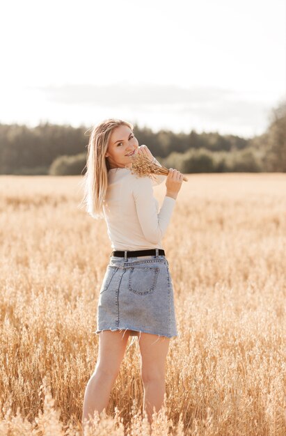 Молодая красивая девушка в джинсовой юбке идет по пшеничному полю в солнечный день
