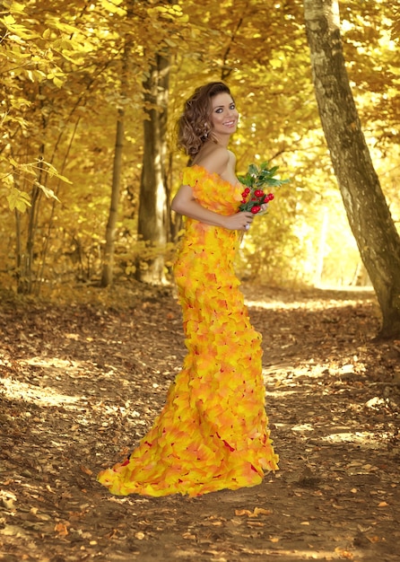 Young beautiful girl in a dress made of autumn leaves in the park in autumn season. Art photo.