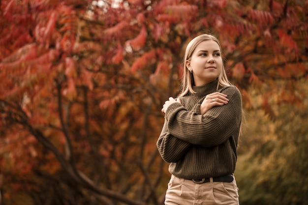 아름다운 나무가 있는 가을 공원에서 세련된 옷, 녹색 스웨터, 베이지색 바지를 입은 젊은 아름다운 소녀