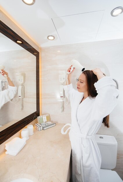 Молодая красивая девушка сушит волосы феном в красивой белой ванной комнате. Свежее доброе утро в отеле.