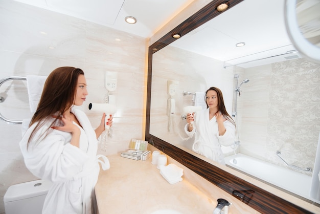 Foto una giovane bella ragazza si asciuga i capelli con un asciugacapelli in un bellissimo bagno bianco