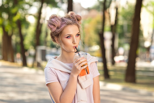 Young beautiful girl drinking fresh juice from plastic takeaway food cups after a walk outdoors. Healthy lifestyle. Smiling slim blonde with pink hair.