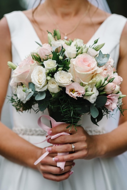 A young beautiful girl in an elegant dress is standing and holding a hand bouquet of flowers and greens with ribbon at nature The bride holds a wedding bouquet outdoors Close up