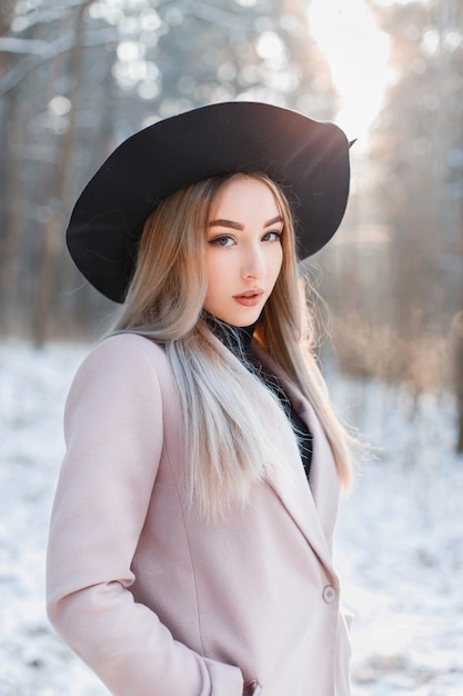 Young beautiful girl in fashionable stylish dress standing in a snowy winter park at sunset