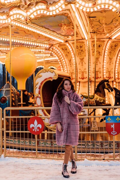 A young beautiful girl in a fur coat stands near the carousel on a winter evening