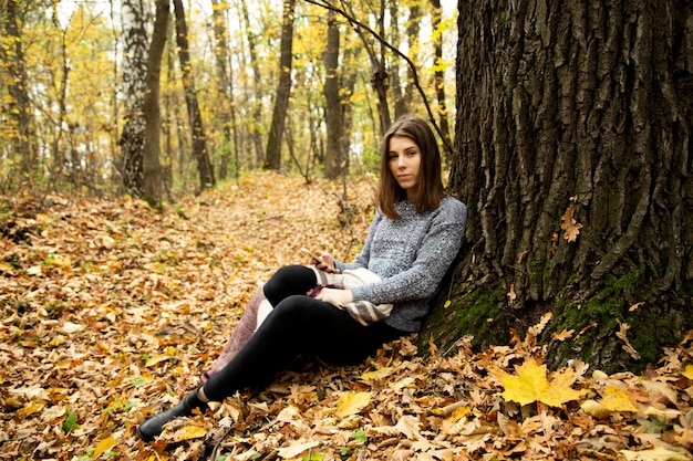 Young beautiful girl in a gray jacket sitting in the autumn forest near a large tree with a mobile phone