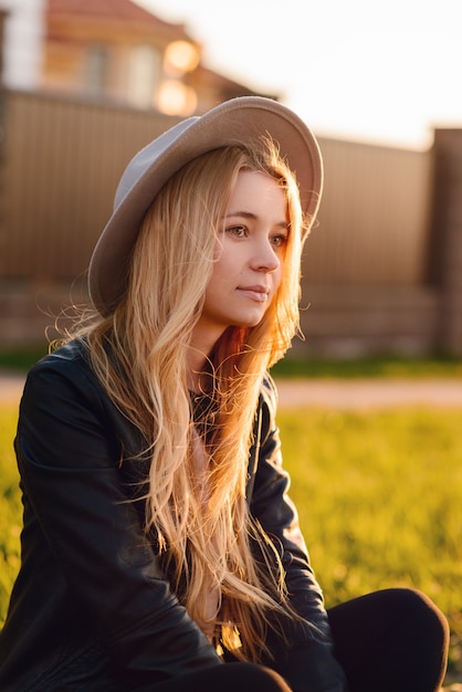 Молодая и красивая девушка в шляпе сидит на обочине дороги на закате