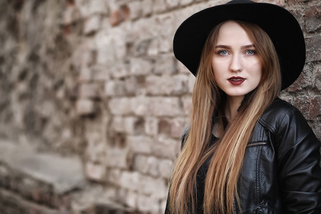 Young beautiful girl in a hat and with a dark make-up outside. Girl in the Gothic style on the street. A girl walks down the city street in a leather waistcoat with phone.