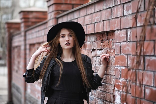 Young beautiful girl in a hat and with a dark make-up outside. Girl in the Gothic style on the street. A girl walks down the city street in a leather waistcoat with phone.