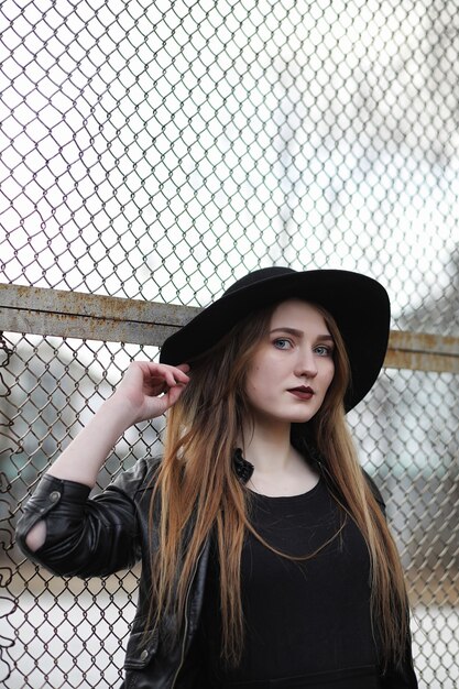 Giovane bella ragazza con un cappello e con un trucco scuro all'esterno. ragazza in stile gotico per strada. una ragazza cammina per la strada della città in un gilet di pelle con il telefono.