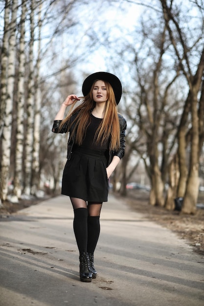 Giovane bella ragazza con un cappello e con un trucco scuro all'esterno. ragazza in stile gotico per strada. una ragazza cammina per la strada della città in un gilet di pelle con il telefono.