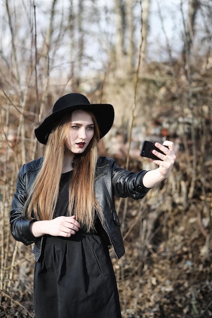 Young beautiful girl in a hat and with a dark makeup outside Girl in the Gothic style on the street A girl walks down the city street in a leather waistcoat with phonexA